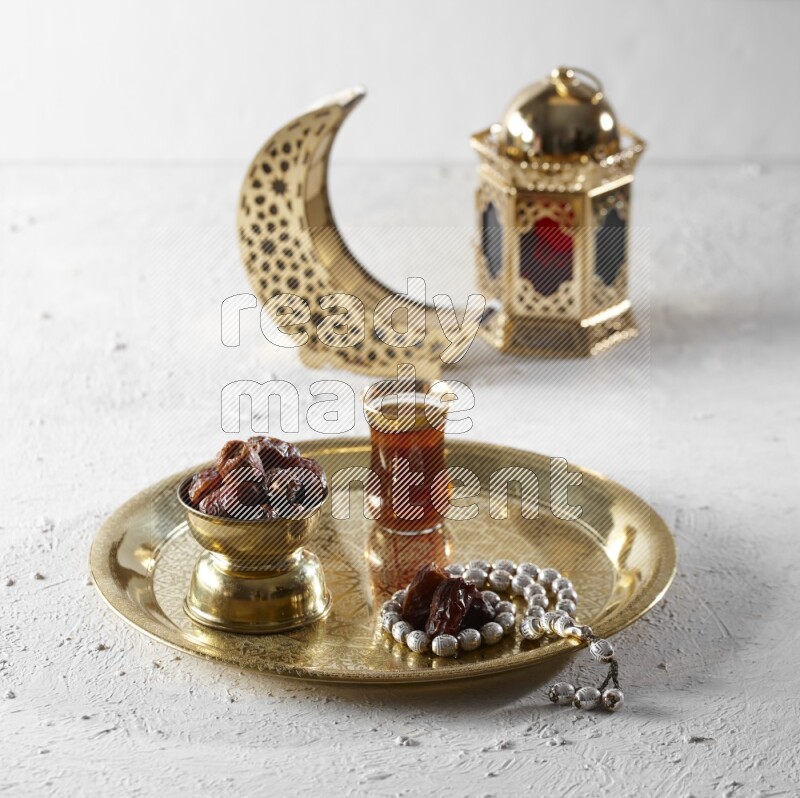 Dates in a metal bowl with tea and prayer beads on a tray beside lanterns in a light setup