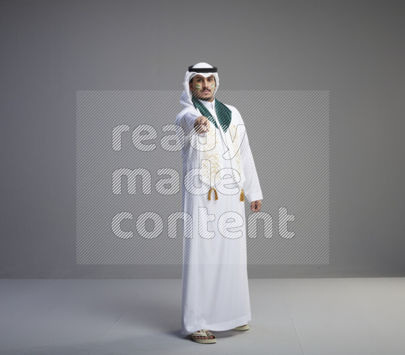 A Saudi man standing wearing thob and white shomag with face painting and Saudi flag scarf on gray background