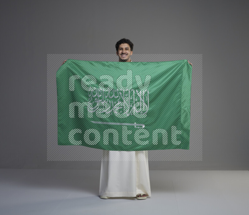 A Saudi man standing wearing thob holding big Saudi flag on gray background