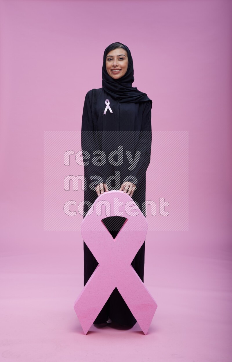 Saudi woman wearing pink ribbon on abaya standing holding awareness ribbon on plain pink background