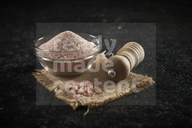 A glass bowl full of pink himalayan salt with a wooden grinder on a burlap fabric all on black background