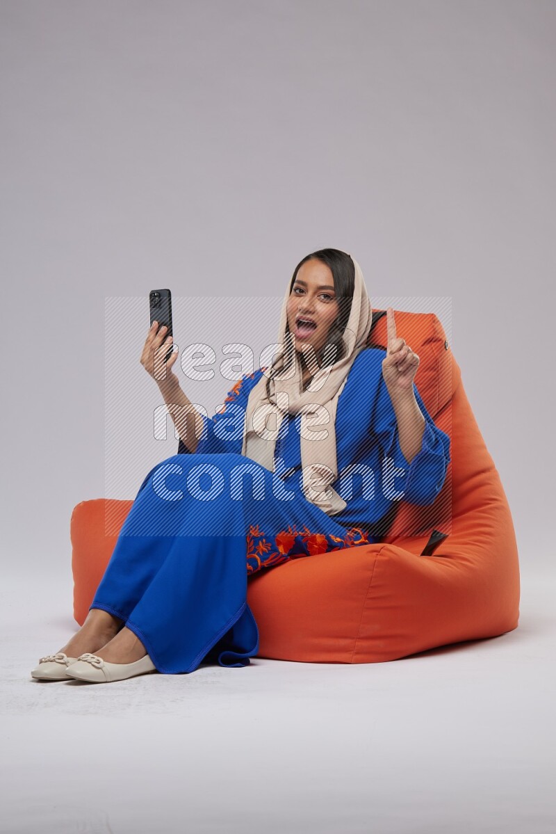 A Saudi woman wearing jalabiya sitting on an orange beanbag and taking selfie