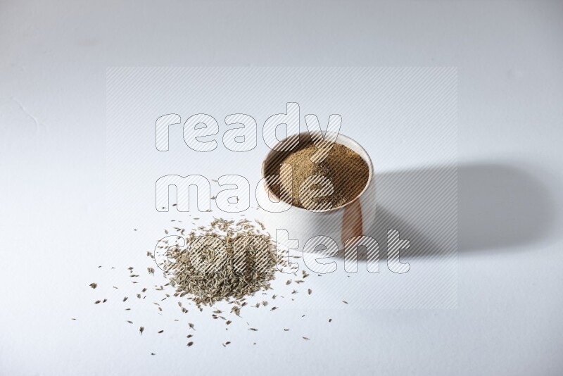 A beige bowl full of cumin powder with cumin seeds beneath it the bowl on a white flooring