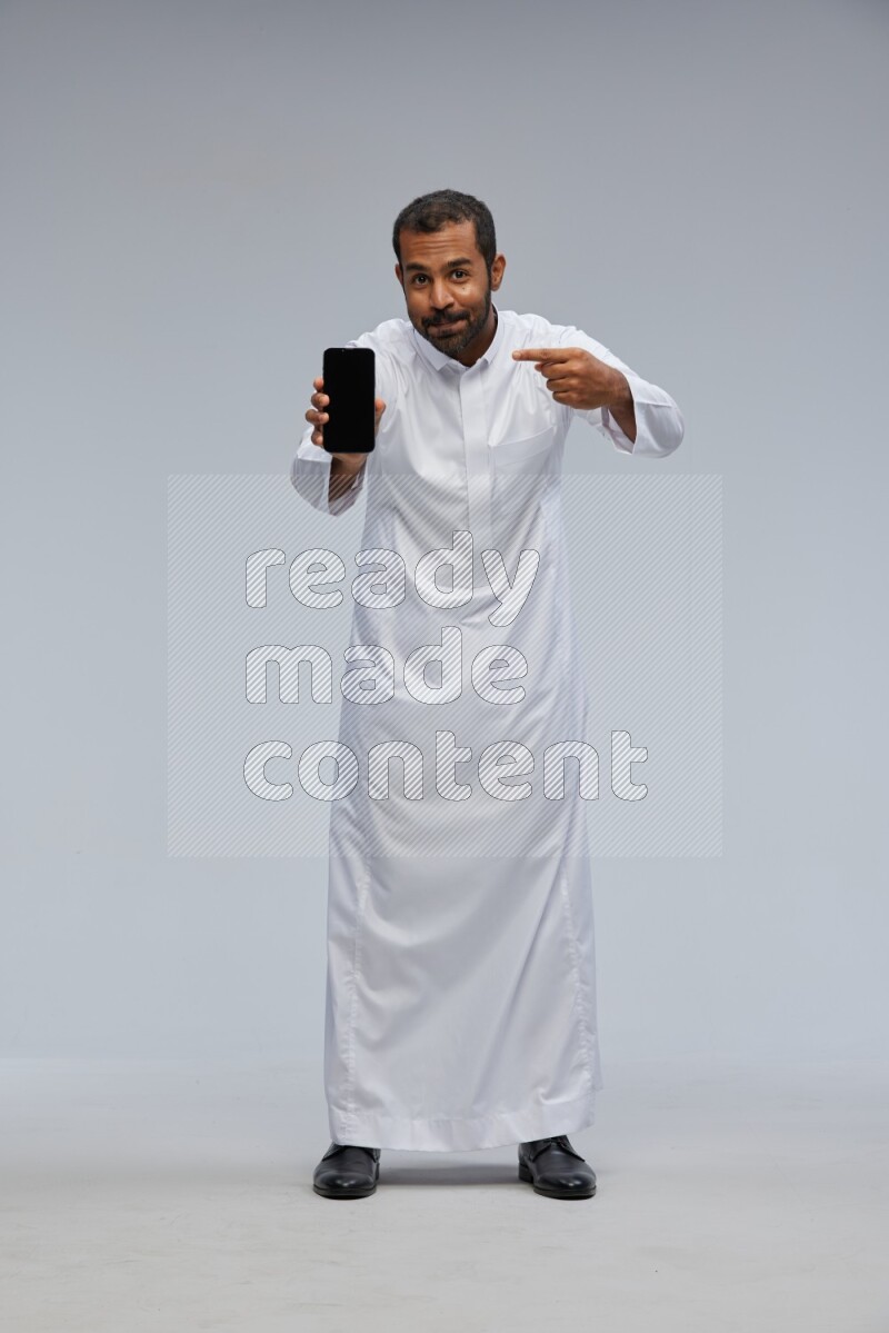Saudi man wearing Thob standing showing phone to camera on Gray background
