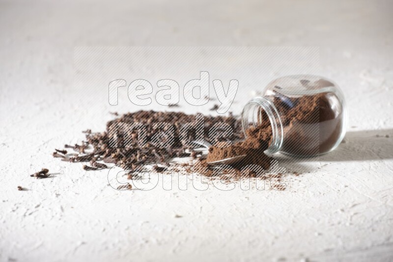 A flipped glass spice jar and a metal spoon full of cloves powder and powder came out of the jar with cloves spread on textured white flooring