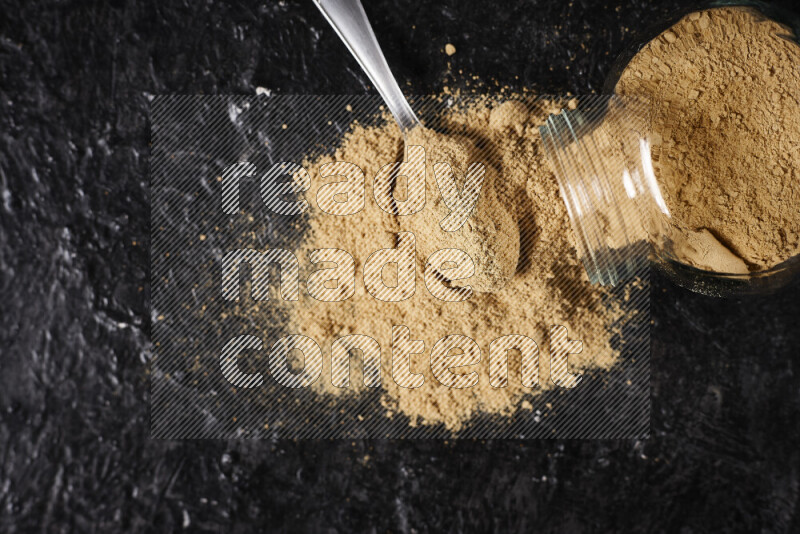 A glass jar full of ground ginger powder flipped with some spilling powder on black background