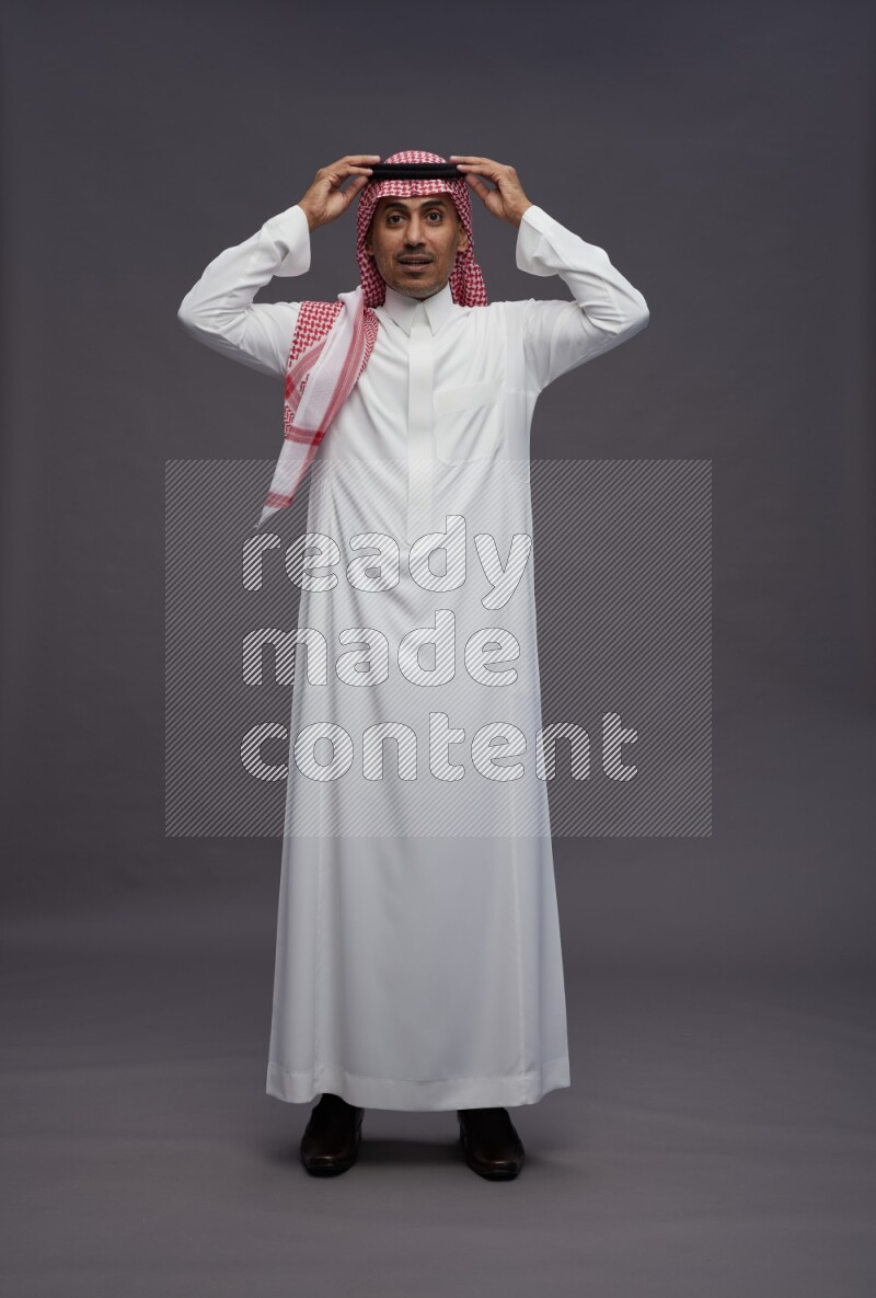 Saudi man wearing thob and shomag standing hands behind head on gray background