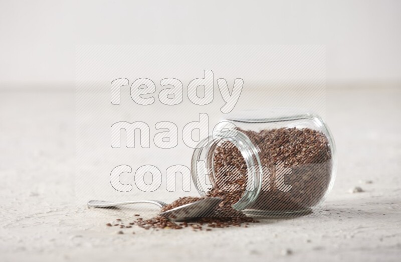 A glass spice jar full of flax seeds flipped with a metal spoon full of the seeds on a textured white flooring