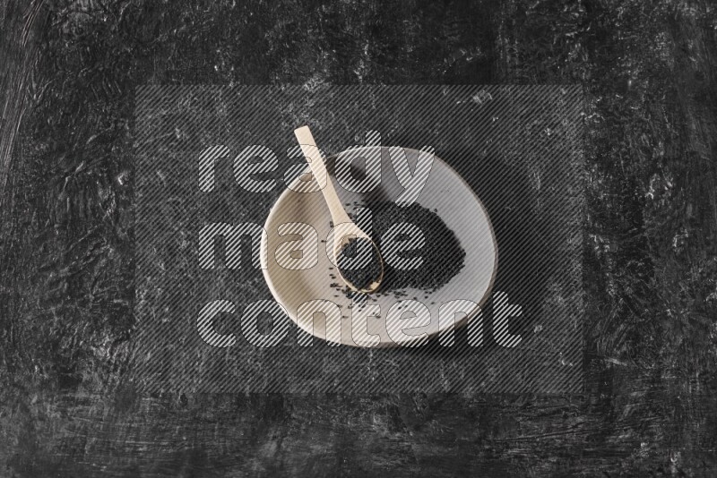 A multicolored pottery plate full of black seeds and wooden spoon full of seeds on a textured black flooring