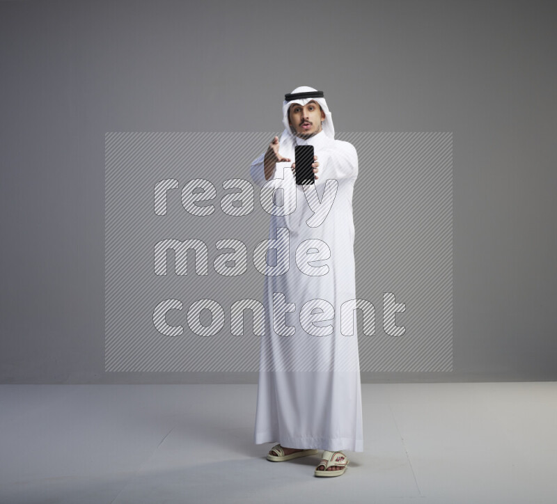 A Saudi man standing wearing thob and white shomag showing phone to camera on gray background