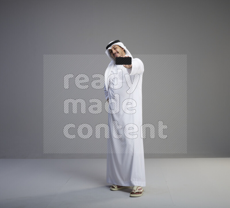 A Saudi man standing wearing thob and white shomag showing phone to camera on gray background