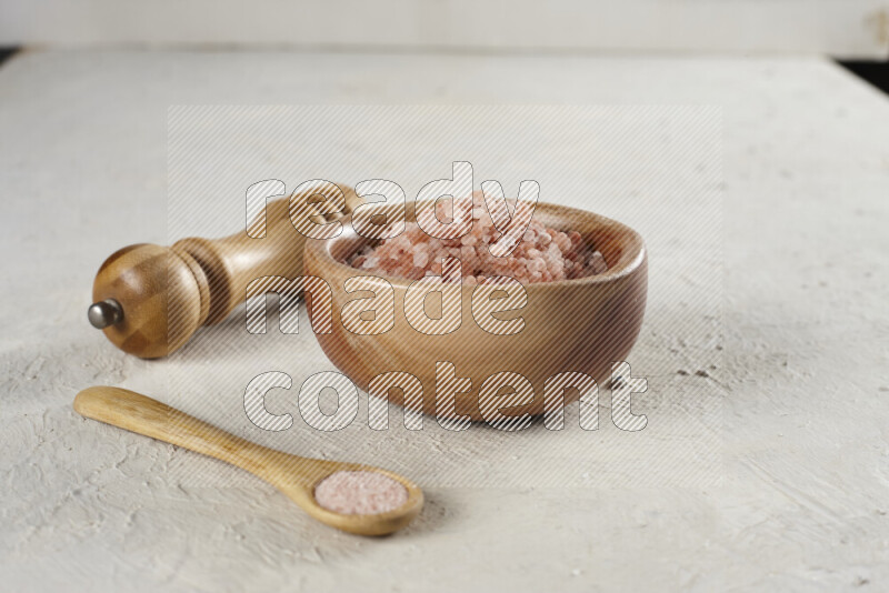 A wooden bowl and spoon filled with coarse pink himalayan salt and a wooden grinder beside them on white background