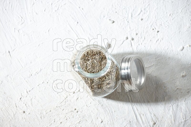 A glass spice jar full of cumin seeds on textured white flooring
