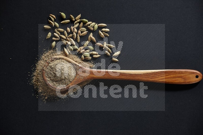 A wooden ladle full of cardamom powder and cardamom seeds beside it on black flooring