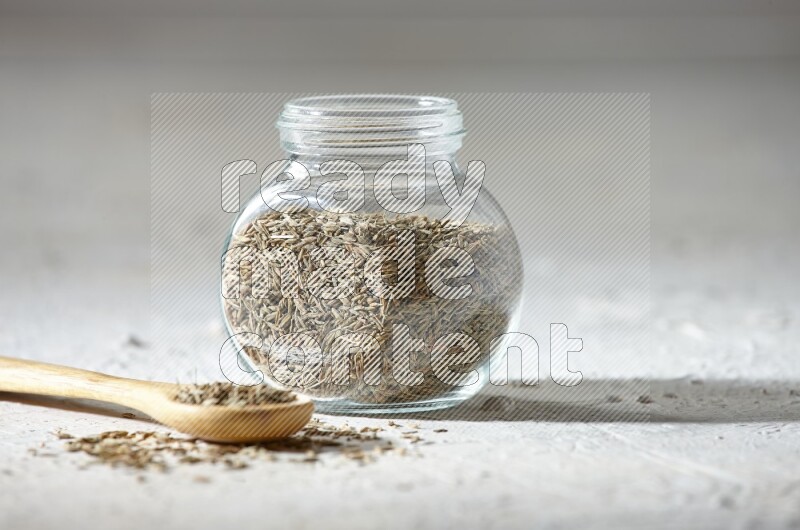 A glass spice jar and wooden spoon full of cumin seeds on textured white flooring