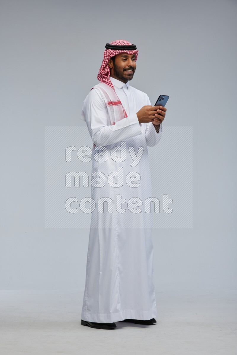 Saudi man Wearing Thob and shomag standing texting on phone on Gray background