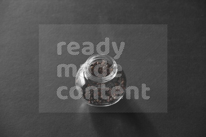 A glass spice jar full of black peppers on black flooring