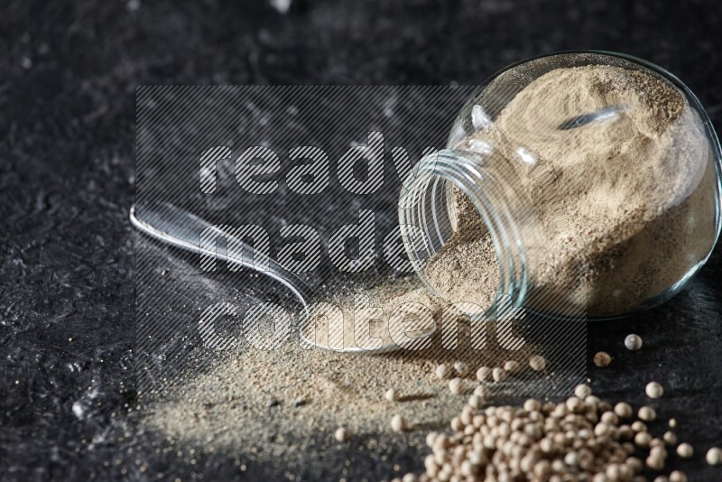 A flipped herbal glass jar and metal spoon full of white pepper powder with spilled powder and pepper beads on textured black flooring