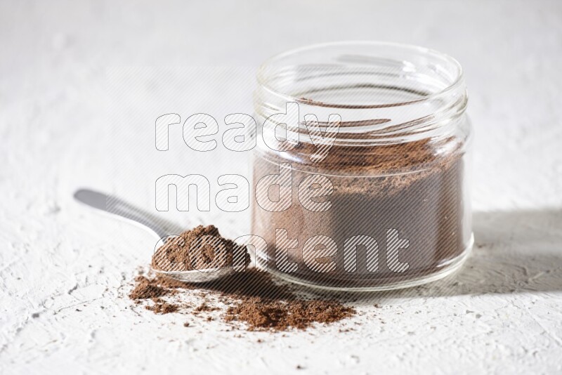 A glass jar full of cloves powder with a metal spoon on a textured white flooring