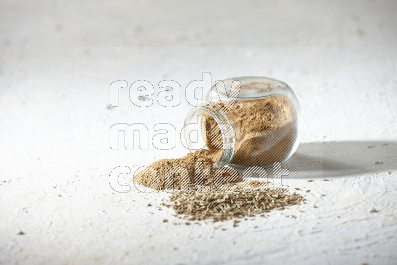 A flipped glass spice jar full of cumin powder and powder spilled out with cumin seeds on textured white flooring