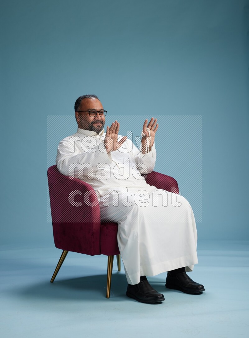 Saudi Man without shimag sitting on chair Interacting with the camera on blue background