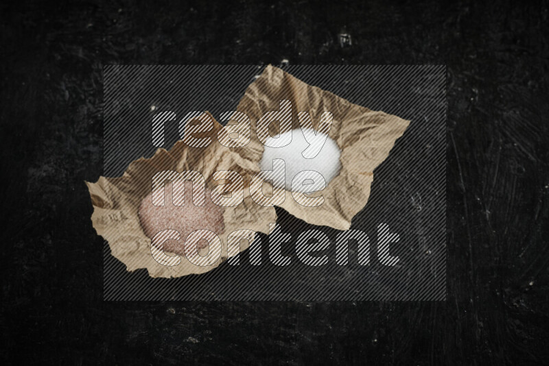 2 crumpled pieces of paper full of pink himalayan salt and white salt on black background