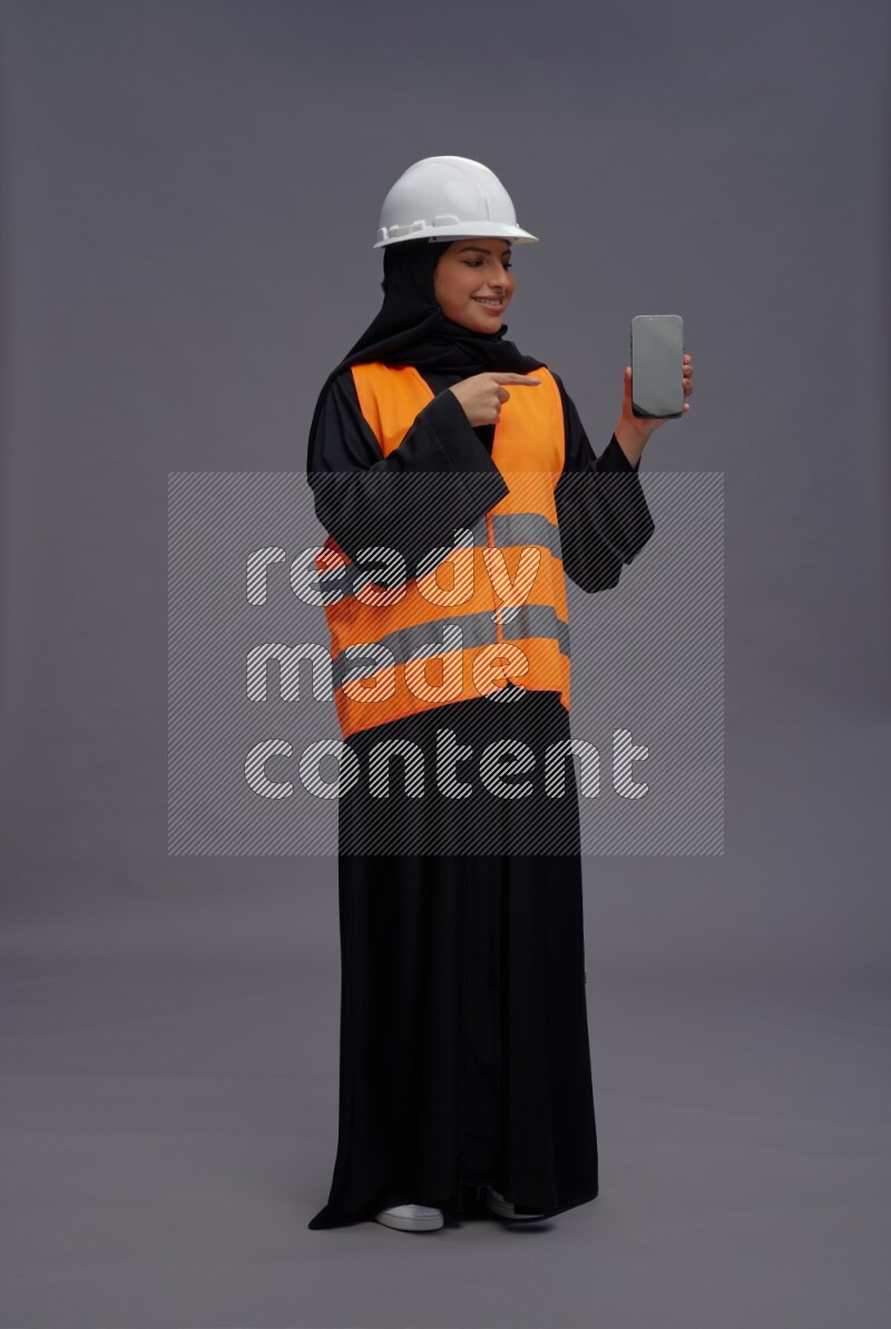 Saudi woman wearing Abaya with engineer vest standing showing phone to camera on gray background
