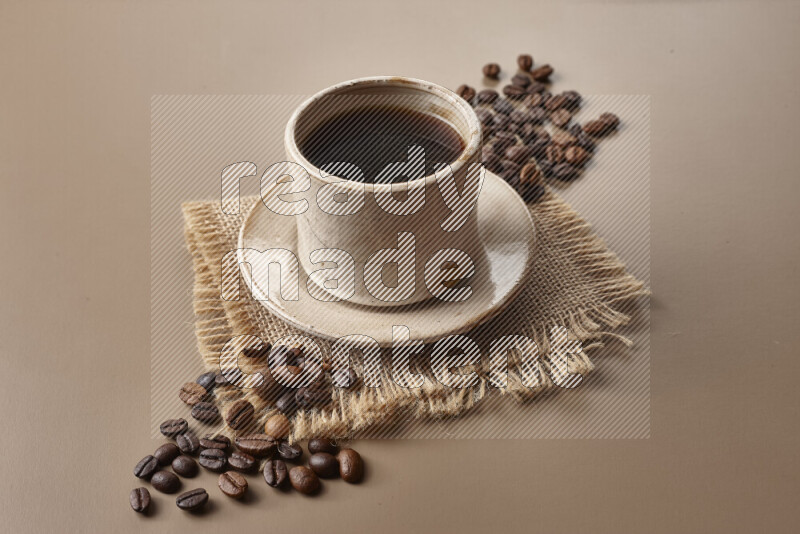 A beige pottery cup of coffee surrounded by roasted coffee beans on beige background