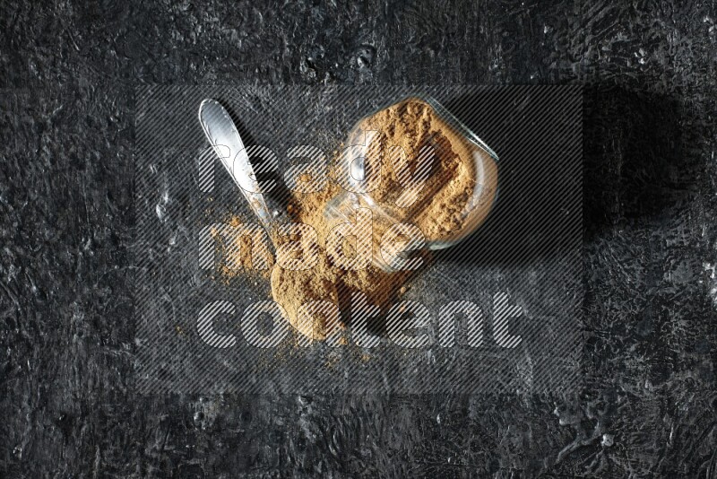 A flipped glass spice jar and a metal spoon full of allspice powder and powder spilled out of it on a textured black flooring