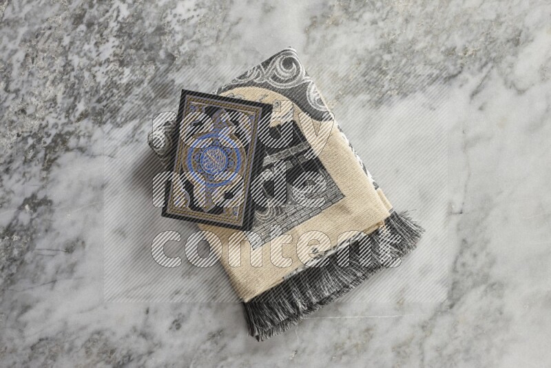 A folded prayer mat with different elements such as quran and prayer beads on grey marble background