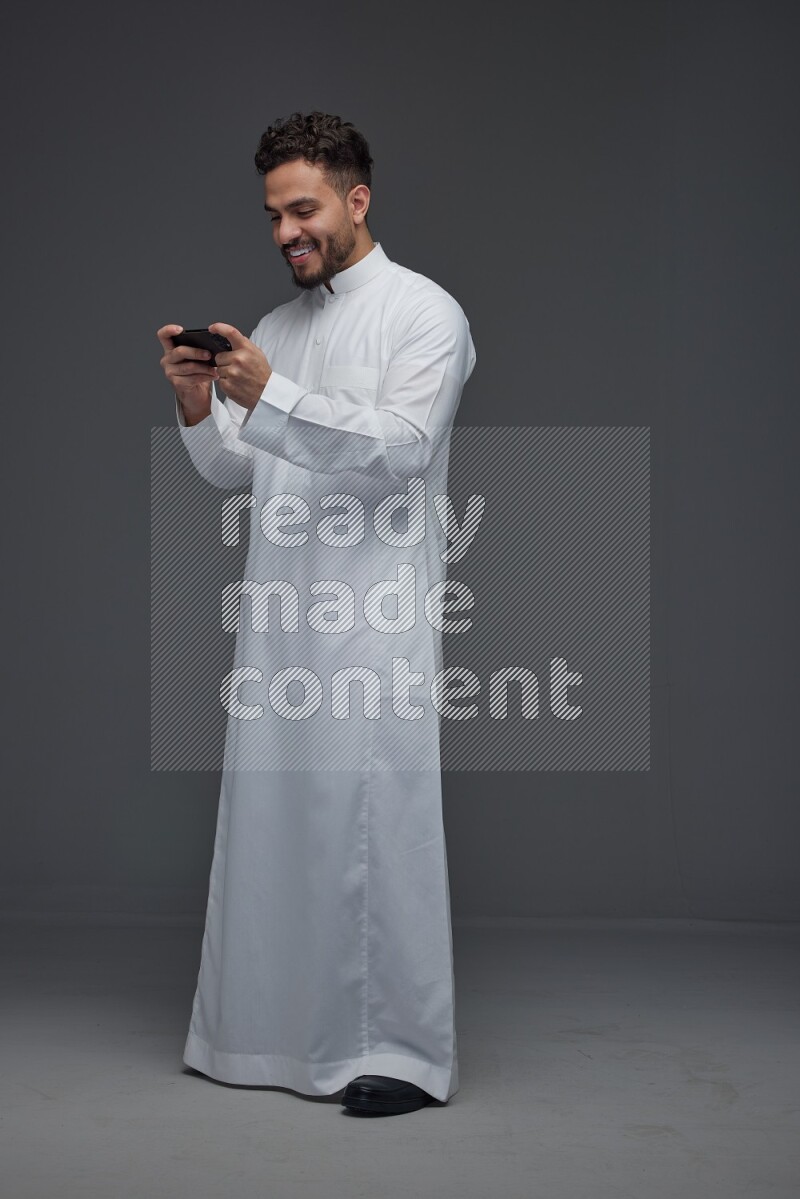 A Saudi man wearing Thobe and playing using his phone horizontally playing video games while standing and making different poses eye level on a gray background