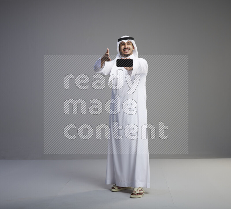 A Saudi man standing wearing thob and white shomag showing phone to camera on gray background