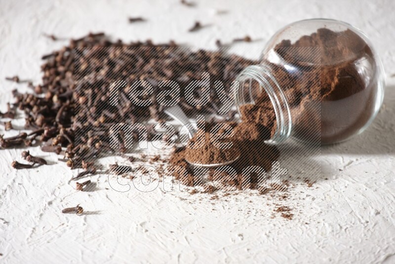 A flipped glass spice jar and a metal spoon full of cloves powder and powder came out of the jar with cloves spread on textured white flooring