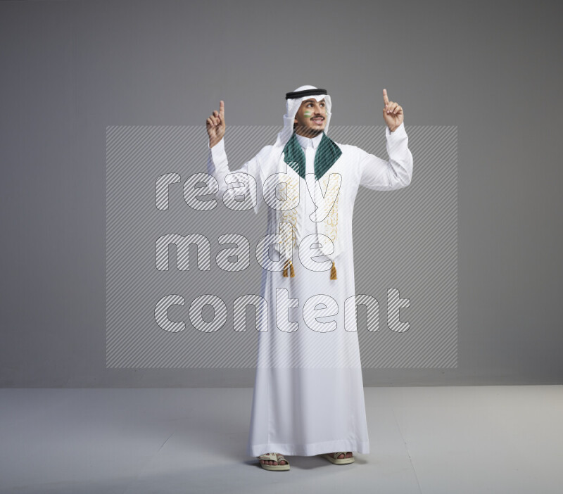 A Saudi man standing wearing thob and white shomag with face painting and Saudi flag scarf on gray background