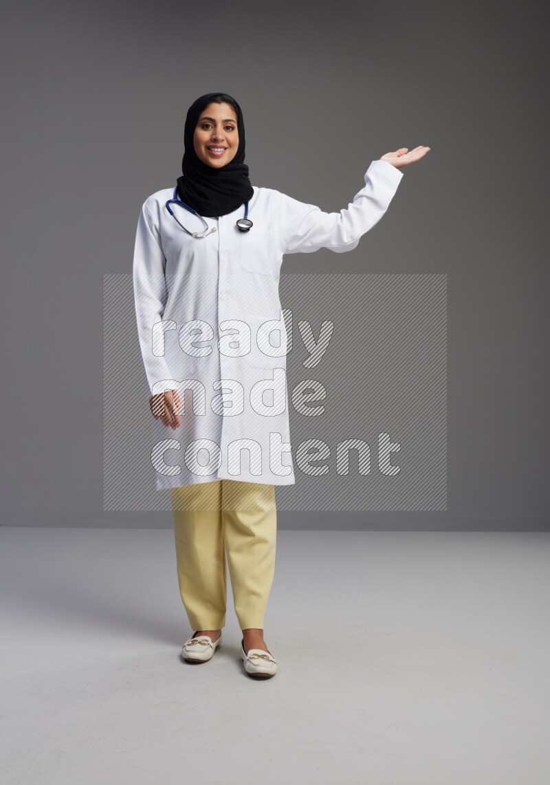 Saudi woman wearing lab coat with stethoscope standing interacting with the camera on Gray background