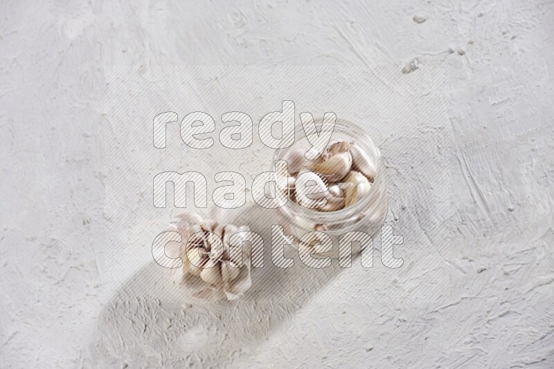 A glass jar full of garlic cloves with a whole garlic bulb beside it on a textured white flooring