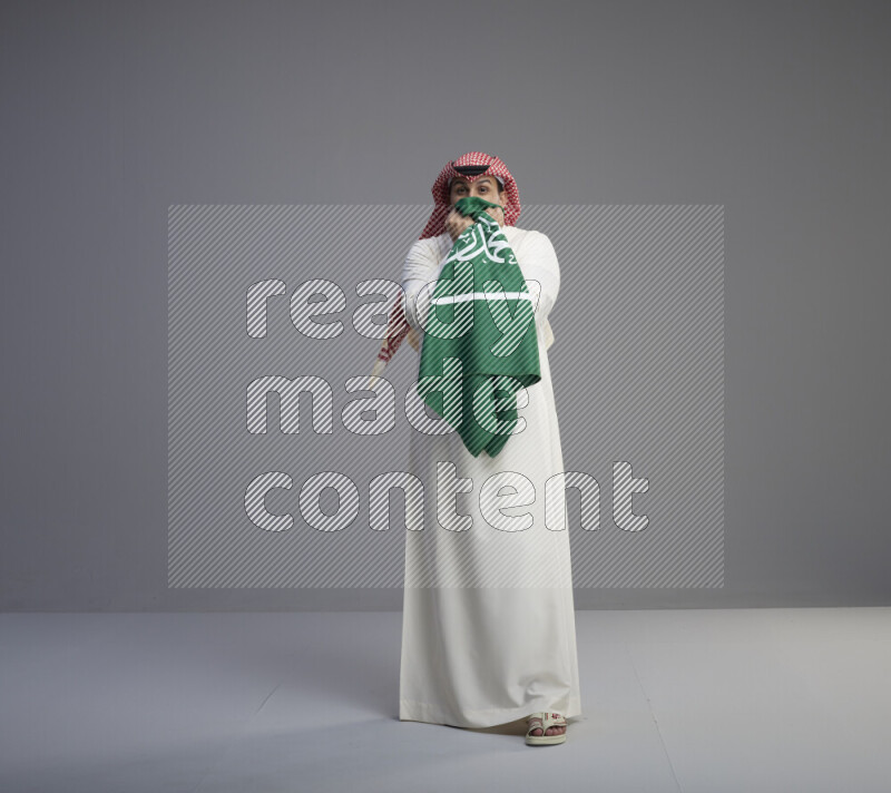 A Saudi man standing wearing thob and red shomag kissing big Saudi flag on gray background