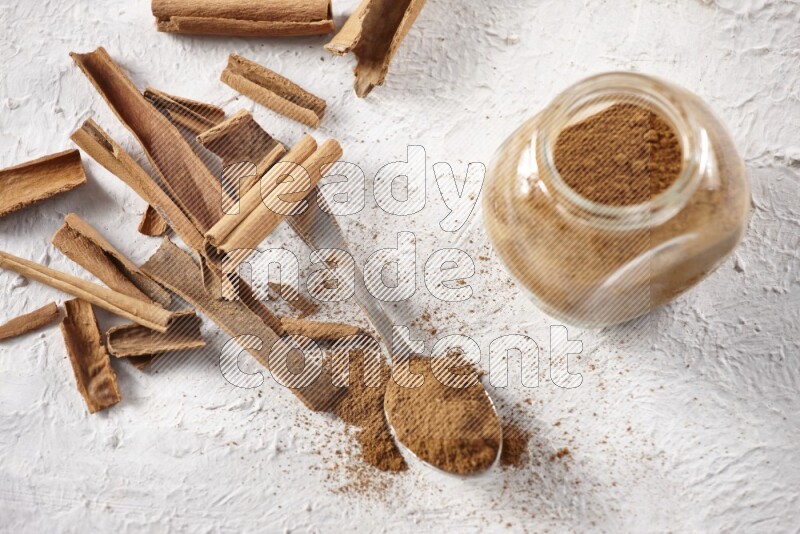 Herbal glass jar full cinnamon powder and a metal spoon surrounded by cinnamon sticks on a white background