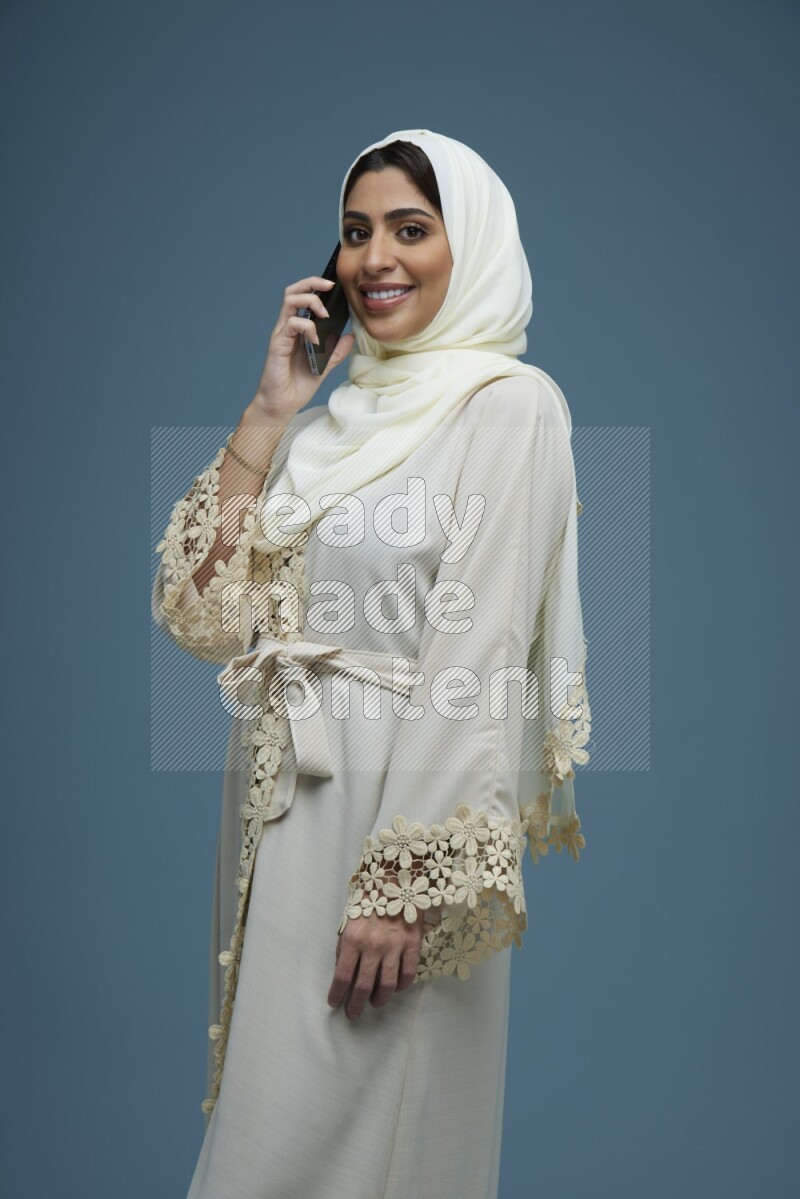 A Saudi woman having a call in a blue background wearing an off-white Abaya Hijab