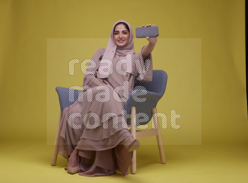 A woman Showing Her Phone Screen Setting  on a Yellow Background wearing Brown Abaya with Hijab