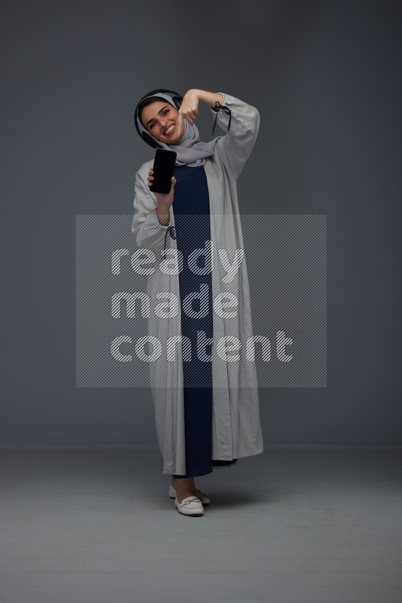 A Saudi woman wearing a light gray Abaya and head scarf setting on a dark grey chair and watching on the tablet eye level on a grey background