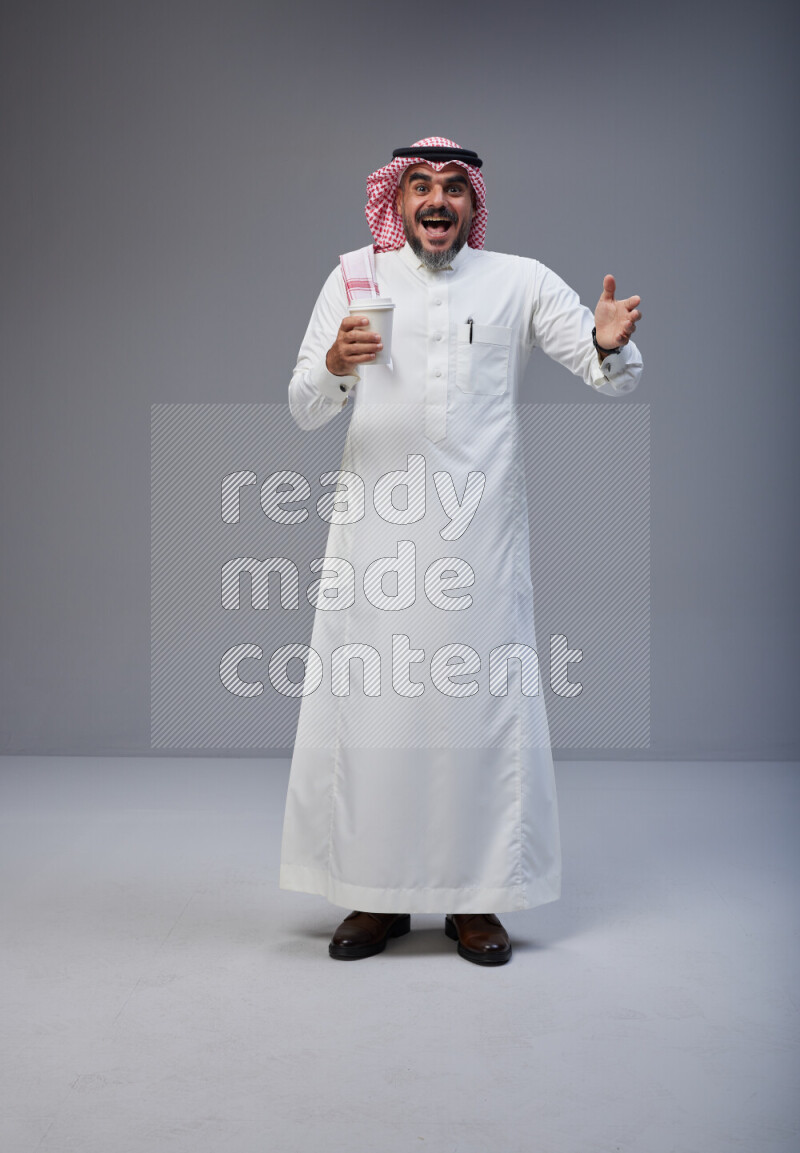 Saudi man Wearing Thob and red Shomag standing holding paper cup on Gray background