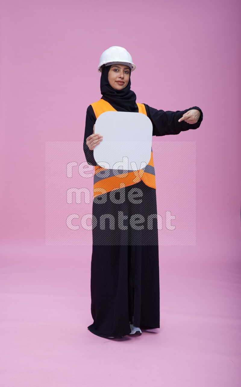 Saudi woman wearing Abaya with engineer vest and helmet standing holding social media sign on pink background