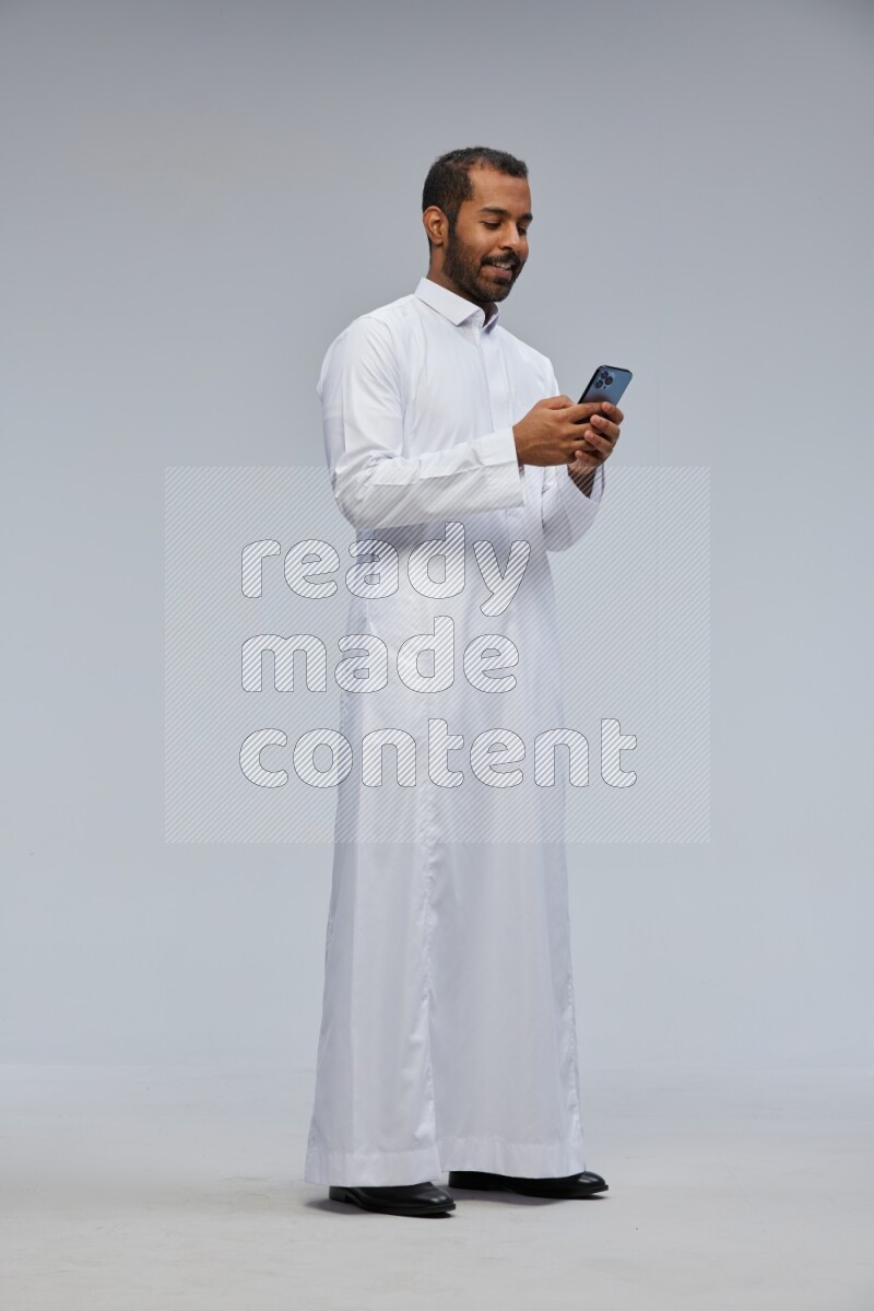 Saudi man wearing Thob standing texting on phone on Gray background