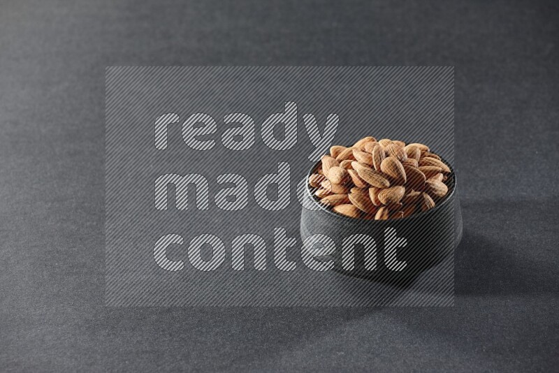 A black pottery bowl full of peeled almonds on a black background in different angles