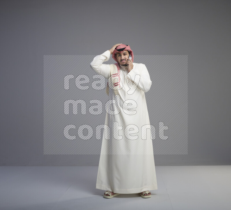 A Saudi man standing wearing thob and red shomag talking on phone on gray background