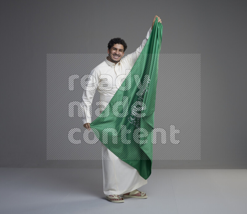 A Saudi man standing wearing thob holding big Saudi flag on gray background