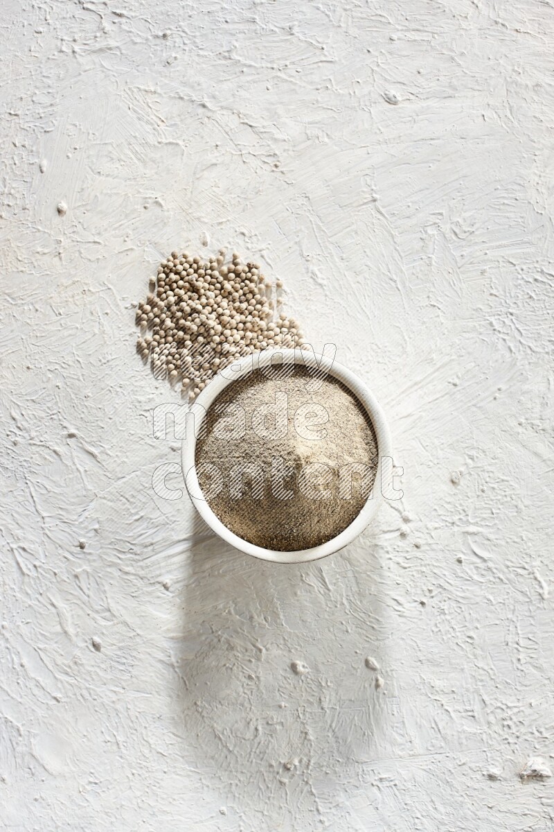 white pottery bowl full of white pepper powder with pepper beads on textured white flooring