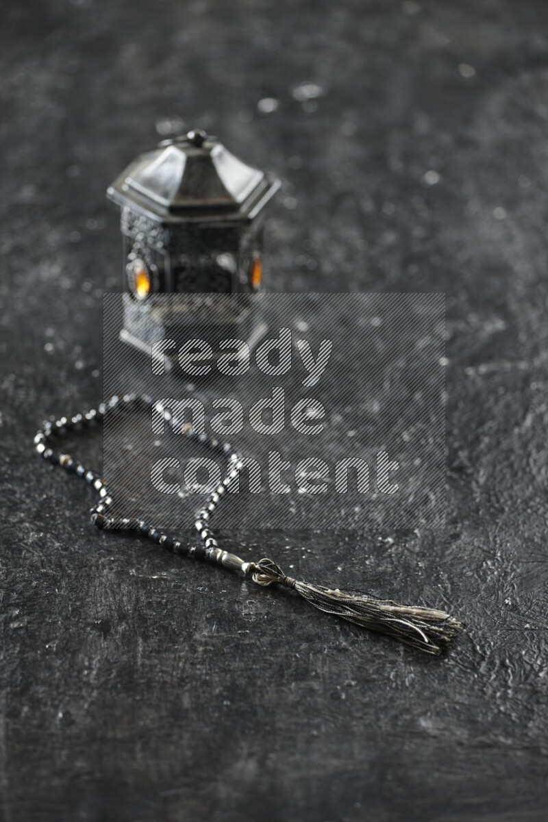A silver lantern with different drinks, dates, nuts, prayer beads and quran on textured black background