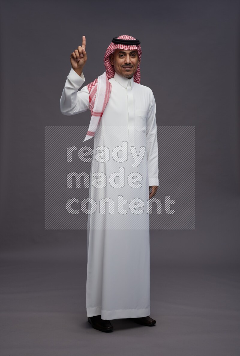 Saudi man wearing thob and shomag standing interacting with the camera on gray background